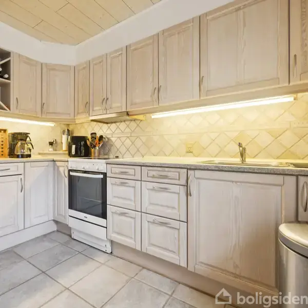 Et køkken med lyse træskabsfronter og flisegulv. En komfur står mellem skabe, over bordpladen med en brødrister og kaffemaskine. Øverst til venstre ses en indbygget vinhylde.