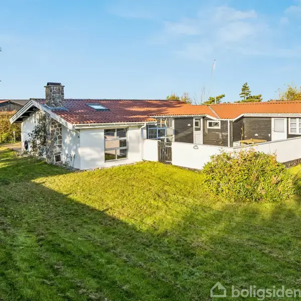 Et hus med rødtag og skorstensrør står i en grøn have. Sollys skinner over græsplænen, og der er buske omkring huset, der er omgivet af træer.