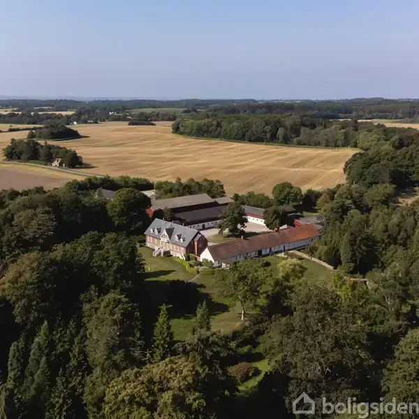 En stor gårdskompleks beliggende i et åbent landskab, omgivet af grønne marker og skovområder under en klar blå himmel.