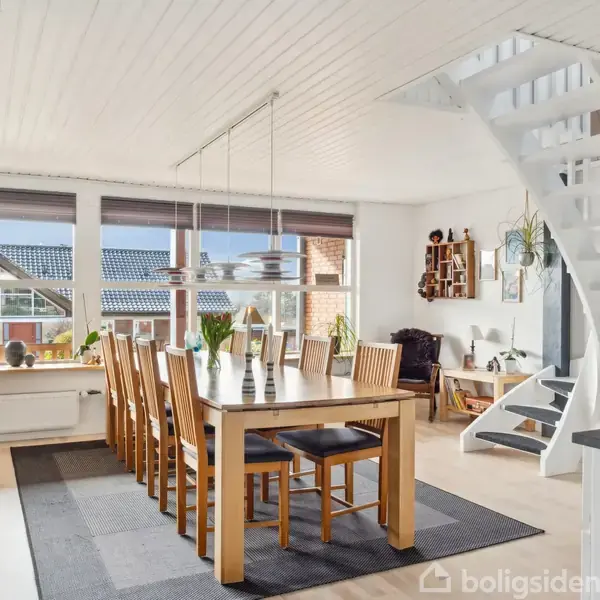 En spisestue med et langt træbord og ti stole står på et tæppe. Store vinduer lader sollys ind, mens en hvid trappe i baggrunden fører til en øvre etage. Planter og hylder pynter rummet.