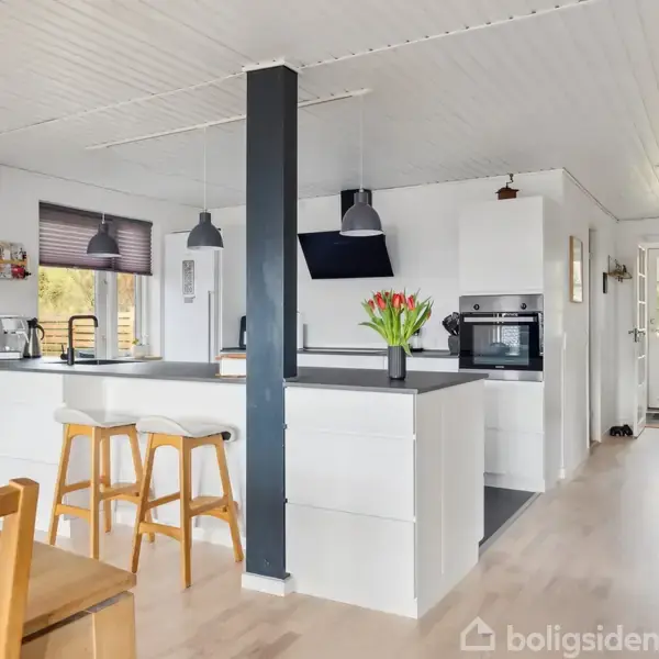 Et spisebord med stole står i et lyst rum med store vinduer, der lader sollys trænge ind. Trappen fører op til en anden etage, omgivet af planter og dekorationer.