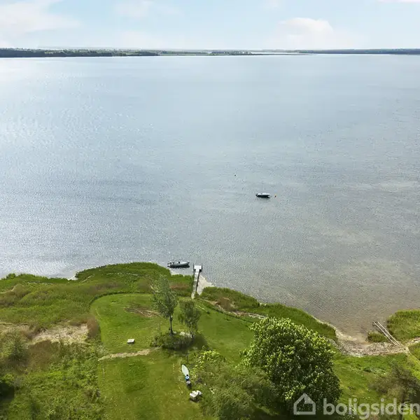En træbro strækker sig ud over en stor sø, mens en lille båd flyder i vandet. Omkringliggende grønne marker og træer skaber en rolig og naturskøn atmosfære.