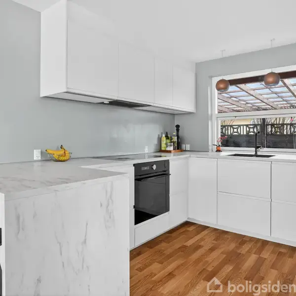 Hvidt, moderne køkken i et hjørneformat. Køkkenet har trægulv, hvide skabe, indbygget sort ovn og marmorlignende bordplade. Et vindue viser en overdækket terrasse udenfor. Der er to sorte barstole.