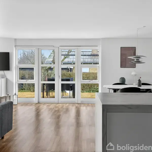 Stue med moderne møbler åbner mod en terrasse gennem glasdøre. Inde i rummet ses et spisebord med stole og væghylder. Udenfor er der en grøn have.