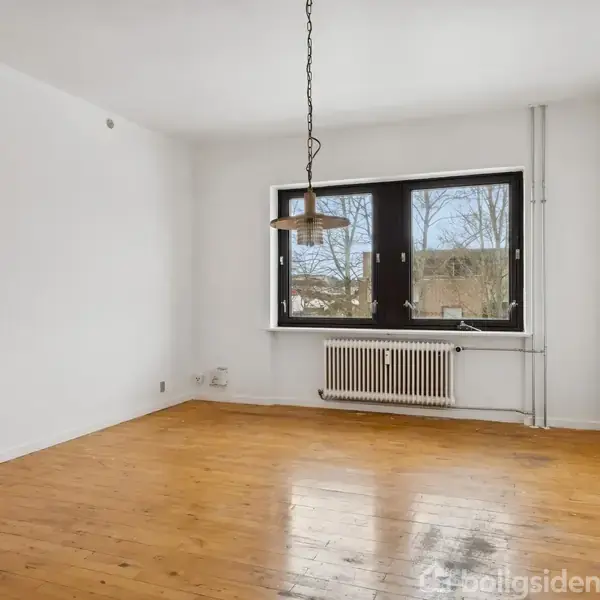 Et tomt værelse med et trægulv, en hængende loftslampe, og et vindue med udsigt til træer udenfor. En radiator er placeret under vinduet.