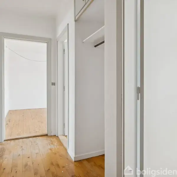 En lys gang med trægulv strækker sig gennem et hus, omgivet af hvide vægge og dørkarme, med åbninger til flere værelser. Tekst “boligsiden” ignoreres.