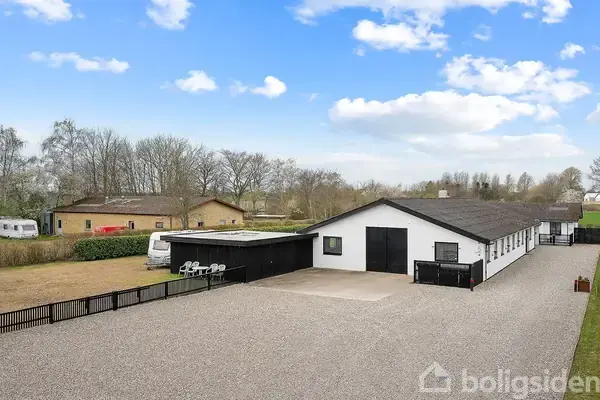 En hvid bygning med sort tag står stille på en grusbelagt gårdsplads omgivet af grønne områder og træer. Der er yderligere mindre bygninger og campingvogne i baggrunden.