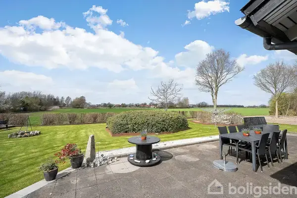 Terrassen har en rund grill og sort spisebord med stole, placeret mod en græsplæne med træer og hække, under en blå himmel med skyer.