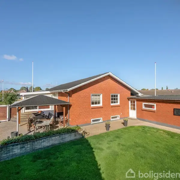 Rødt murstenshus står stille i en solrig baghave med velplejet græsplæne. Der er en flisebelagt terrasse med overdækket siddeområde til venstre. Blå himmel og enkelte skyer i baggrunden.