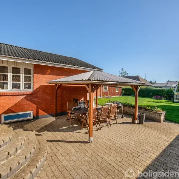 En overdækket terrasse med borde og stole står foran et rødt murstenshus. I baggrunden er der en velplejet græsplæne og træer under en klar blå himmel.