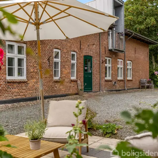 En terrasse med en parasol og havemøbler står foran et murstenshus med hvide vinduer og en grøn dør. Blomster omkranser terrassen; træer ses i baggrunden.