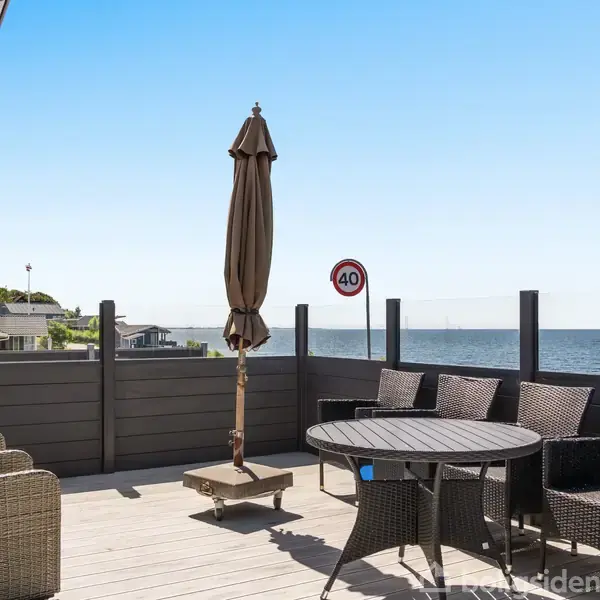 Terrassemøbler er placeret på en balkon med udsigt over havet. Et træbord og fluktstole står i solen. Et skilt viser "40" kilometer i timen.