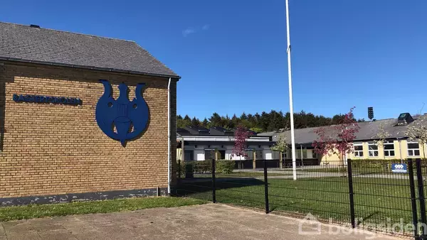 En brun murstensbygning med et blåt skjold symbol på væggen. Tekst: "ROSENKILDESKOLEN". Bygningen er omgivet af grønne områder med et metalhegn og en flagstang, under en blå himmel.