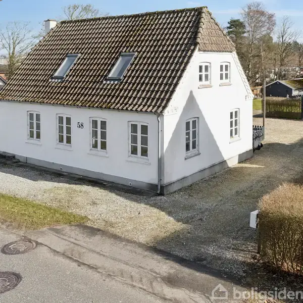 Hus med hvid facade og brunt tag står stille ved en vej med nummeret 58 tydeligt synligt. Omkringliggende have med indkørsel og små drivhuse.