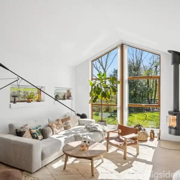 Stue med hvid sofa og træbord, oplyst af naturligt lys fra store vinduer. En sort brændeovn står ved siden af. Grønne planter og kunstværker fuldender det lyse, moderne look.