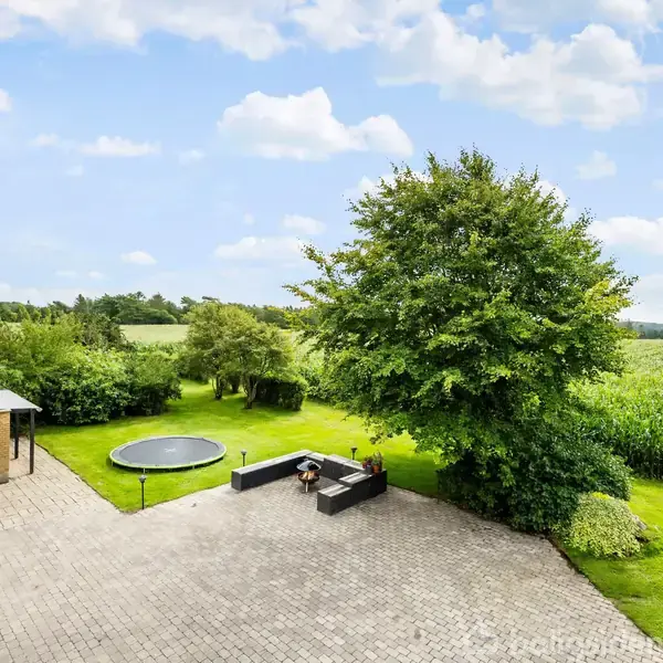 Trampolin i gårdhave omgivet af grønne områder og træer ved siden af en bygning med rødt tag; mark ses i baggrunden.