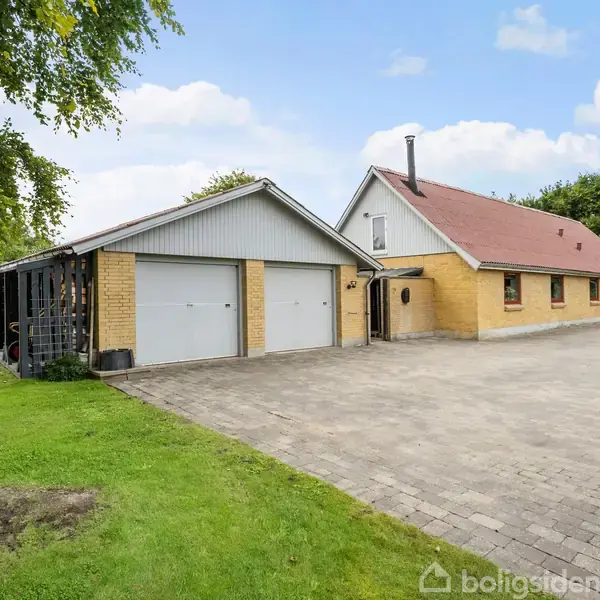 Et gult murstenshus med rødt tag og to hvide garageporte står på en stor, flisebelagt gårdsplads. Omgivet af grønne træer og plæne under en klar blå himmel.