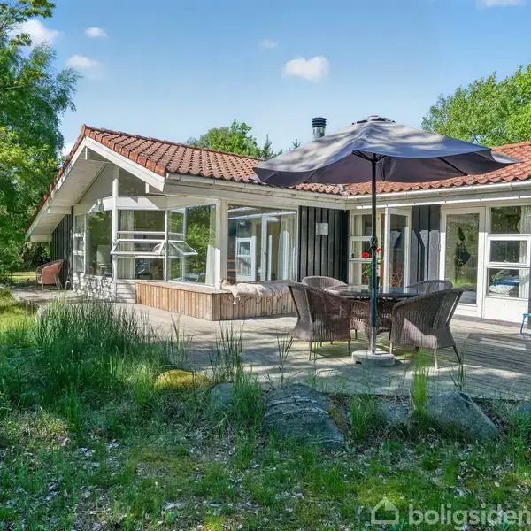 Et sommerhus med rødt tegltag og store vinduer ses. Havemøbler med parasol står på terrassen foran huset. Omgivet af frodig vegetation og blå himmel.