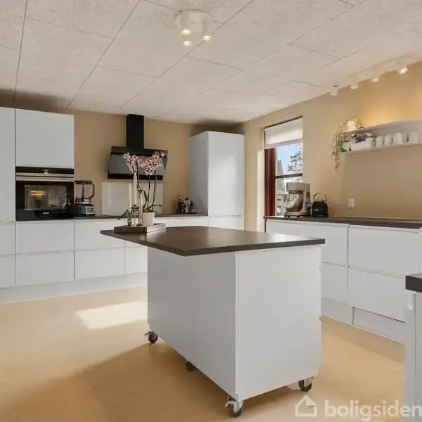 Et moderne køkken med en central ø på hjul, omgivet af hvide skabe. Rumopdelingen er lys, med store vinduer og blomster som dekoration.