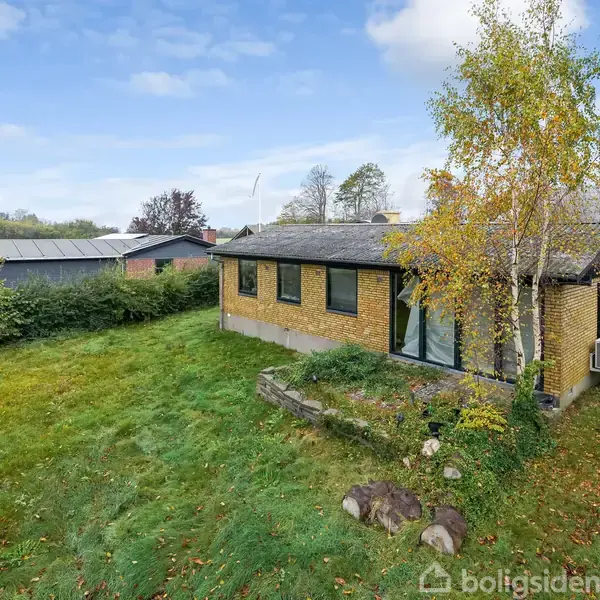 Et gult murstenshus står på en græsplæne omgivet af buske og træer. Der er en stor have med enkelte sten i forgrunden, og flere huse ses i baggrunden.