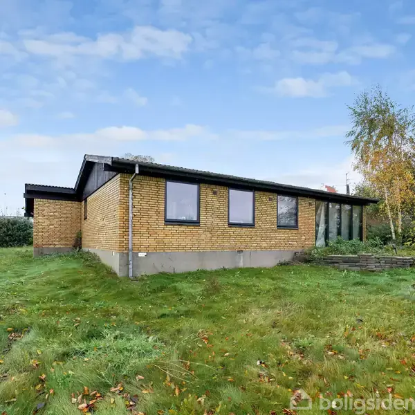 Et gulstenshus står på en græsplæne omgivet af buske og træer. Det har store vinduer og fladt tag under en skyet himmel.
