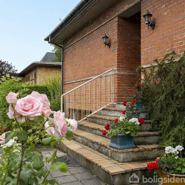 Rosen i forgrunden blomstrer ved siden af en havedekoration. Foran et murstenshus fører en trappe med gelænder op til indgangen. Omkring huset er der grønne planter og blomster.