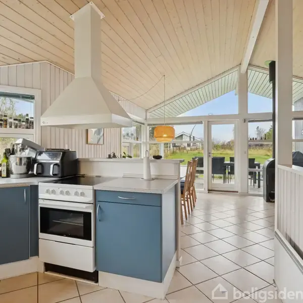 Køkken med komfur og emhætte i moderne stil, omgivet af blågrå skabslåger. I baggrunden ses et spisebord med stole i et rum med store vinduer og udsigt til en have.