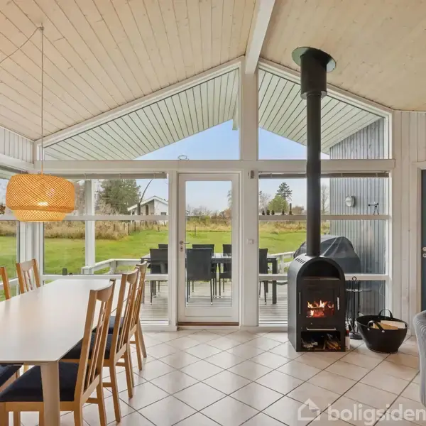 Spisebord med stole står ved vinduer og glasdør. Brændeovn med ild ved siden af. Udenfor ses græsplæne og havemøbler. Hængelampe svæver over bordet.