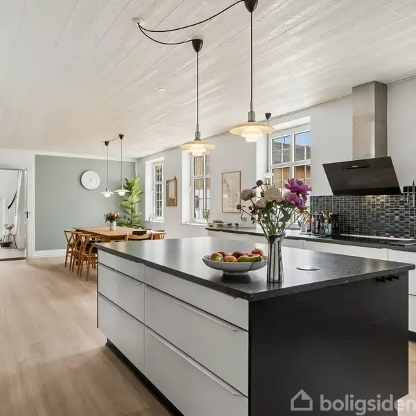 Køkkenø med skål frugt og vase blomster på sort bordplade, omgivet af hvide skabe. Hængelamper ovenover; i baggrunden spisebord og stole i en lys, rummelig stue.
