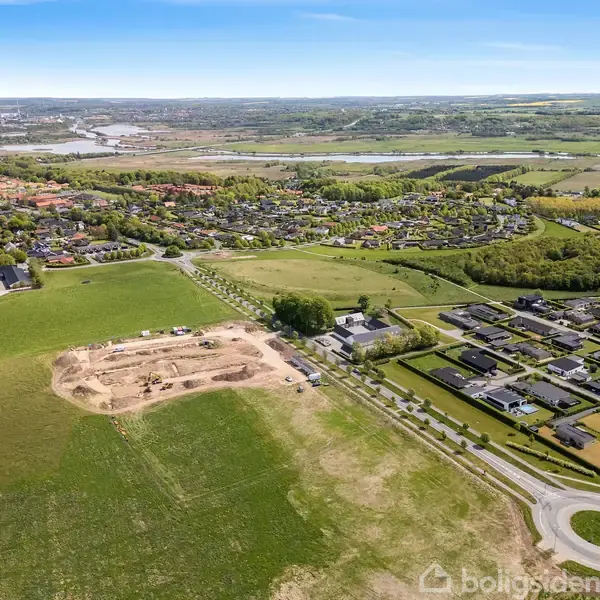 Byggeområde med grusdækning omfavnes af åbne marker og nærliggende boligkompleks. En rundkørsel forbinder veje mellem byggeriet og beboelsesområdet. Floden og byen ses i det fjerne.