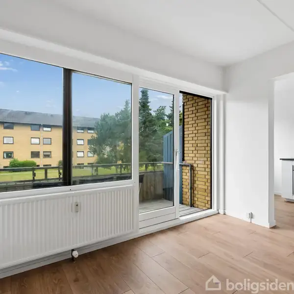 Et lyst rum med store vinduer og en radiator, der åbner ud til en altan med udsigt over en gårdsplads. Ved siden er et køkken med komfur og emhætte.