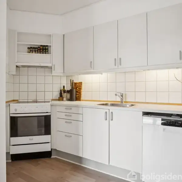 Et moderne køkken med hvide skabe og en indbygget opvaskemaskine. En komfur står til venstre, omgivet af krydderihylder, alt i et lyst rum med flisebeklædte vægge.