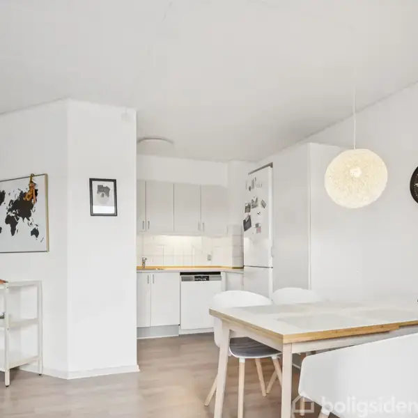 Et lyst køkken-alrum med et træbord og fire hvide stole. Køkkenet har hvide skabe og en opvaskemaskine. En verdenskort plakat hænger på væggen. Klokken viser klokken 10.12.