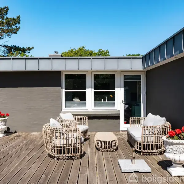 Terrassemøbler står på en trægulvterrasse foran et moderne, enkelt-etagers hus. Plantede blomster i krukker tilføjer farve, og grønne træer ses i baggrunden under en klar blå himmel.