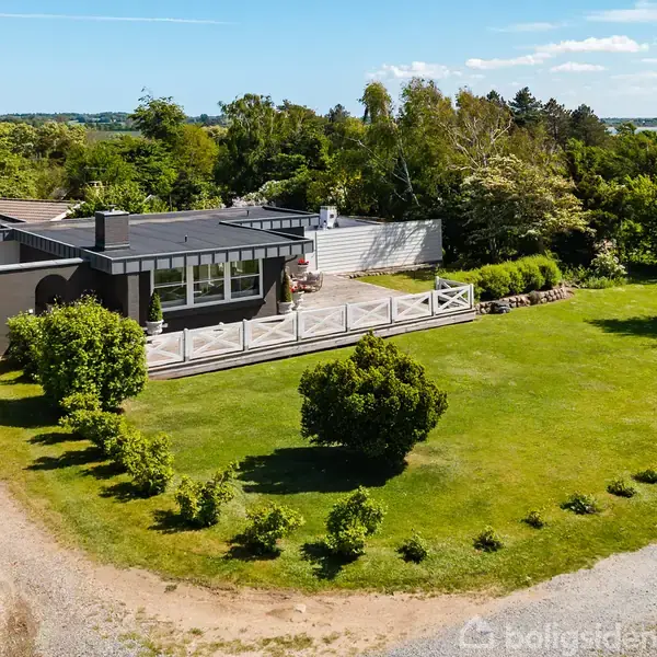 En moderne villa med stor terrasse, beliggende i grønne omgivelser med velplejet have. Huset er sort med hvide detaljer og omgivet af grusveje samt frodig vegetation.