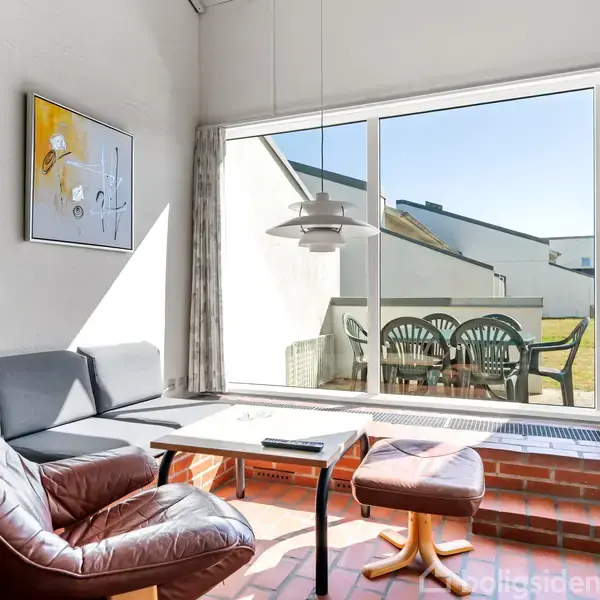 En læderlænestol og sofabord står i en stue med røde klinker og store vinduer, der viser en terrasse med grønne plastikstole udenfor. Et maleri hænger på væggen.