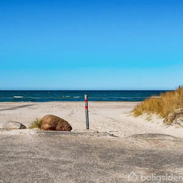 Strand med bløde sandklitter. En hastighedsskilte viser "30". Græs vokser ved klitterne, og havet er roligt i horisonten. Klar blå himmel uden skyer.