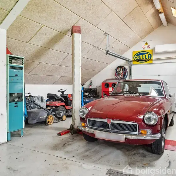 En rød vintagebil holder parkeret på en autolift i en garage. Omkring ses diverse værktøjer, en plæneklipper, og et reklameskilt med teksten "HELLA" på væggen.