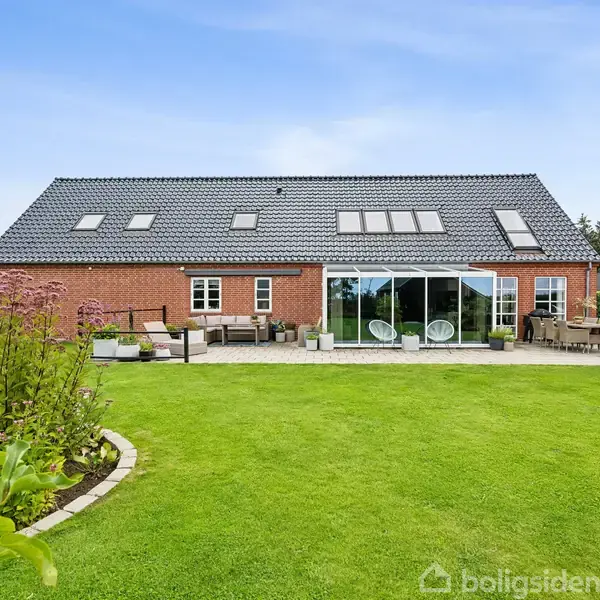 Et rødt murstenshus med mørkt tag har store vinduer. Det står i en velholdt have med grøn græsplæne og terrassemøbler omkring. Skyfri blå himmel i baggrunden.