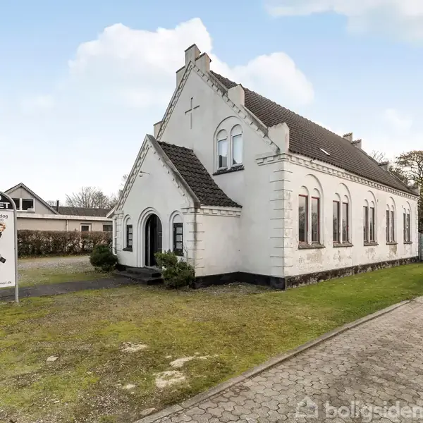 En hvid bygning med mørkt tag står ved en vej. Foran den er et skilt med teksten "SKO OUTLET Jesper Lærke," omgivet af grøn græs og belægningssten.