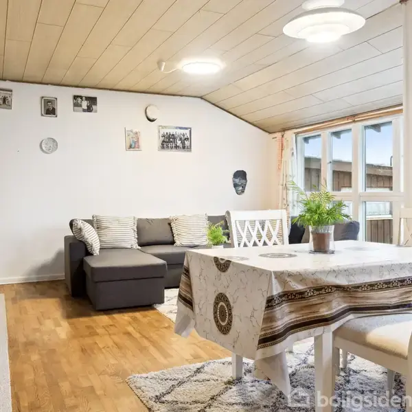 En stue med sofa og bord, som er dækket med en mønstret dug, omgivet af stole. Rummet har træloft og et trægulv, med billeder hængende på væggen og grønne planter i vinduet.