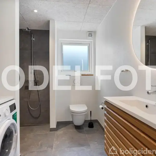 Vaskemaskine står stille ved siden af bruser i moderne badeværelse. Rundt spejl med lys hænger over hvid vask på træbord. VKord "MODELFOTO" ses midt i billedet.