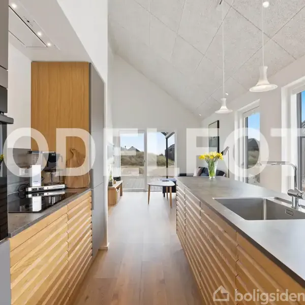 Køkken med moderne indretning, viser en lang køkkenbordplade med indbygget vask. Lyse pendellamper hænger over bordet. Store vinduer giver udsigt til en have. "MODEL FOTO" står skrevet centralt.