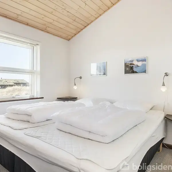 En dobbeltseng med hvide puder og dyner står i et lyst værelse. Væggene er hvide med to små billeder, og loftet er af lyst træ. To lamper hænger ved siden af sengen. En vindue viser udsigt til træer og sand.