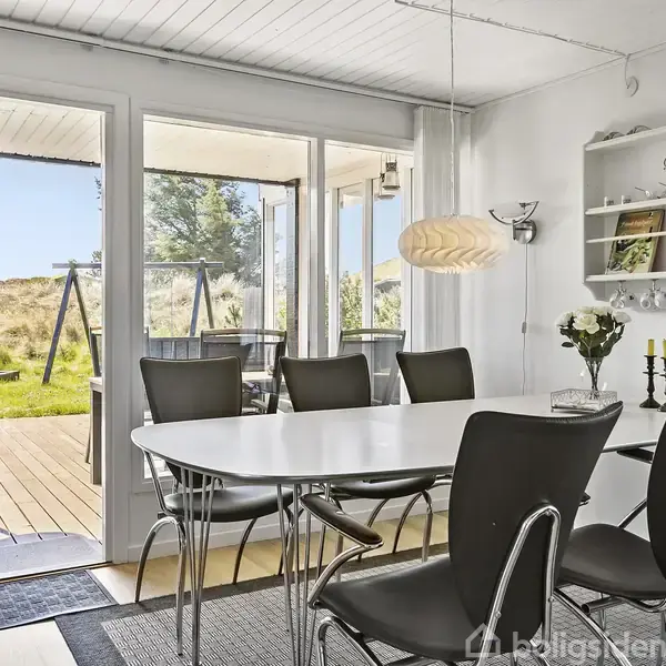 Spisebord med seks sorte stole er omgivet af et lyst rum med åbne terrassedøre. Udenfor ses en græsplæne med gyngestativ og træer i baggrunden.