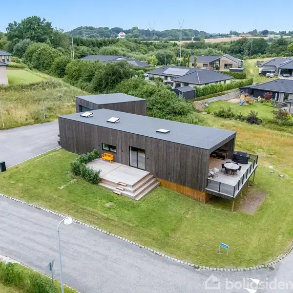 Moderne træhus med fladt tag ligger på hjørnet af boligområde. Foran huset er en terrasse med trappe. Omkringliggende grønne områder og flere andre huse i nabolaget.