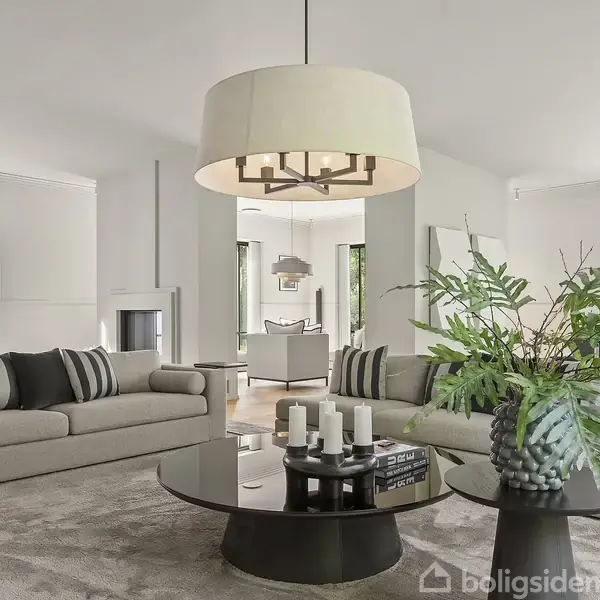 En moderne stue med to grå sofaer omkring et rundt sofabord, der har stearinlys på. Store vinduer slipper lys ind fra haven udenfor.