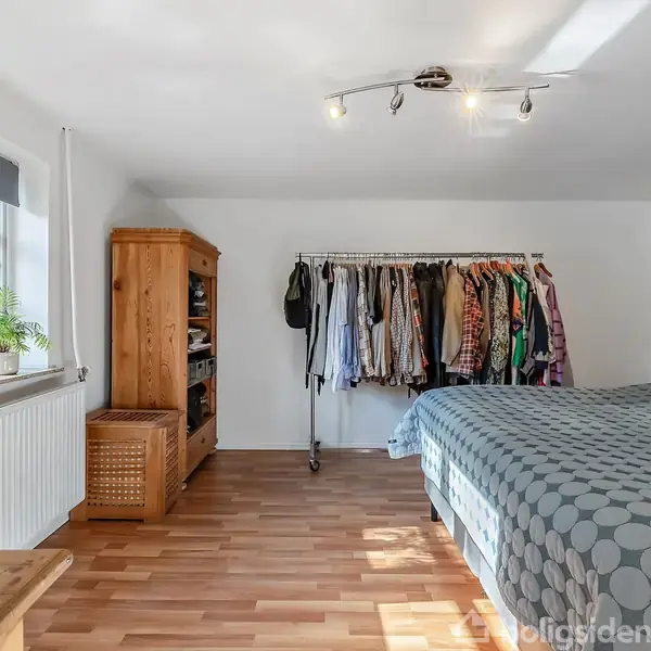 Soveværelse med seng dækket af gråt sengetæppe. Tøj hænger på en garderobestativ. Træskab og kurv ved vindue med planter. Rummet har lyst trægulv og hvidmalede vægge.