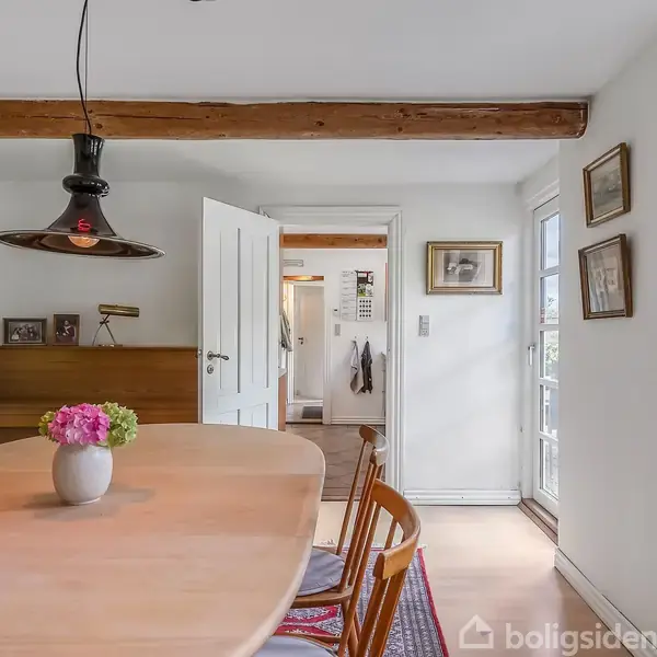 Et spisebord med en vase fyldt med blomster står i et lyst rum. Stole omgiver bordet, og en åben dør leder til et andet rum. Væggene har vinduer og billeder.