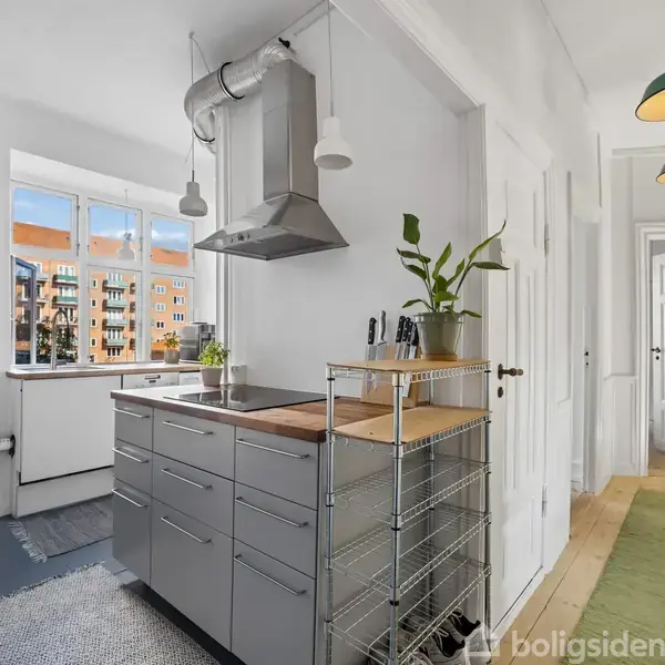 Køkken med moderne gråt køkkenskab, aftræksemhætte, og planter på køkkenø. Store vinduer bringer lys ind, og en smal gang fører videre. Grønne lamper hænger fra loftet.